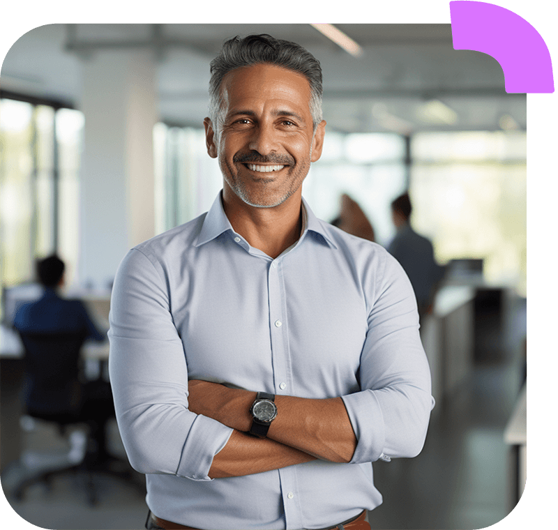 Um homem confiante de meia-idade, com cabelos grisalhos e barba bem aparada, sorrindo de forma calorosa. Ele está vestindo uma camisa social azul claro com as mangas dobradas e tem os braços cruzados. Ele está em um ambiente de escritório moderno, com outras pessoas trabalhando ao fundo.